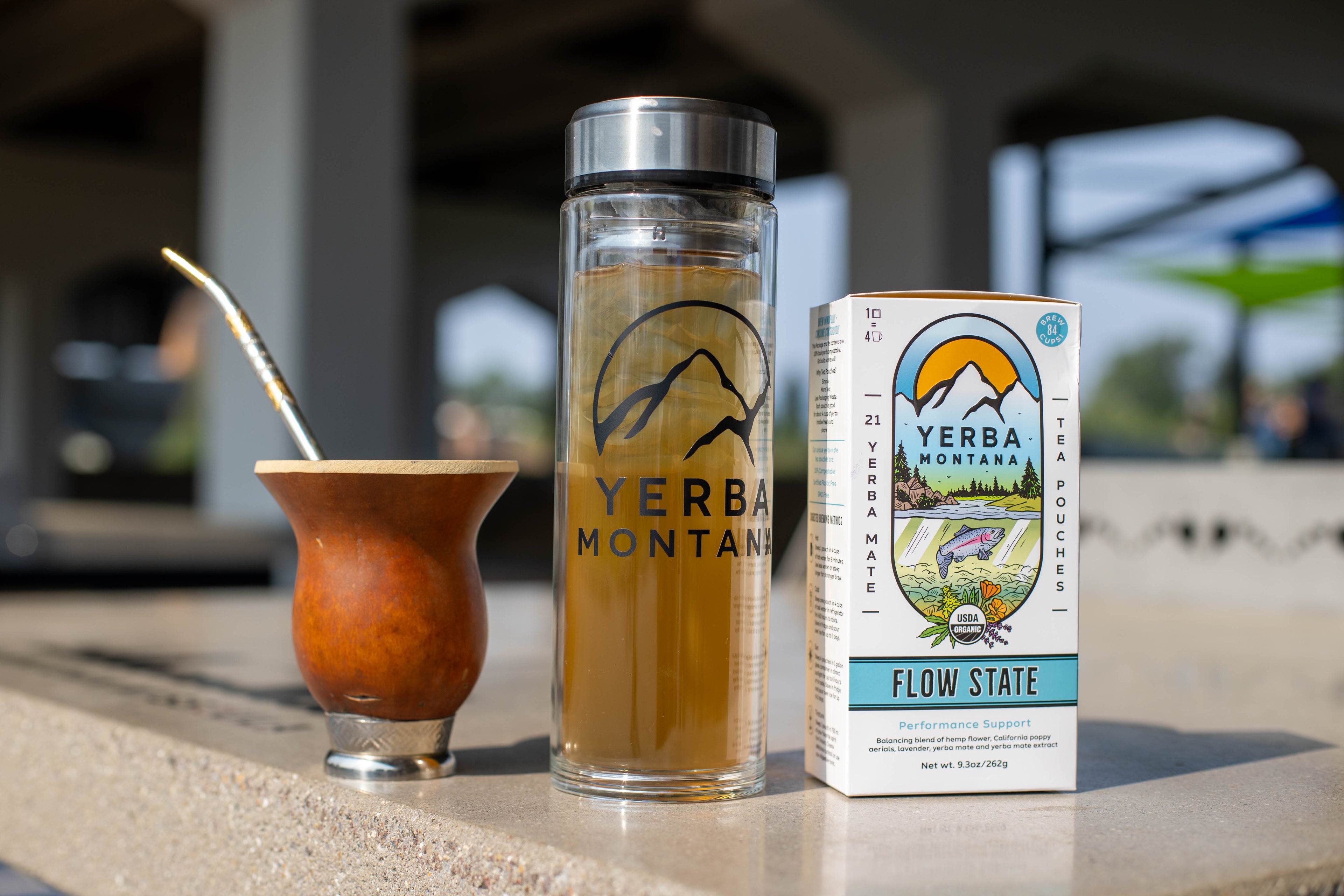 Yerba Montana gourd with bombilla on a stone table, clear thermos of iced mate, and Flow State tea pouch box.

