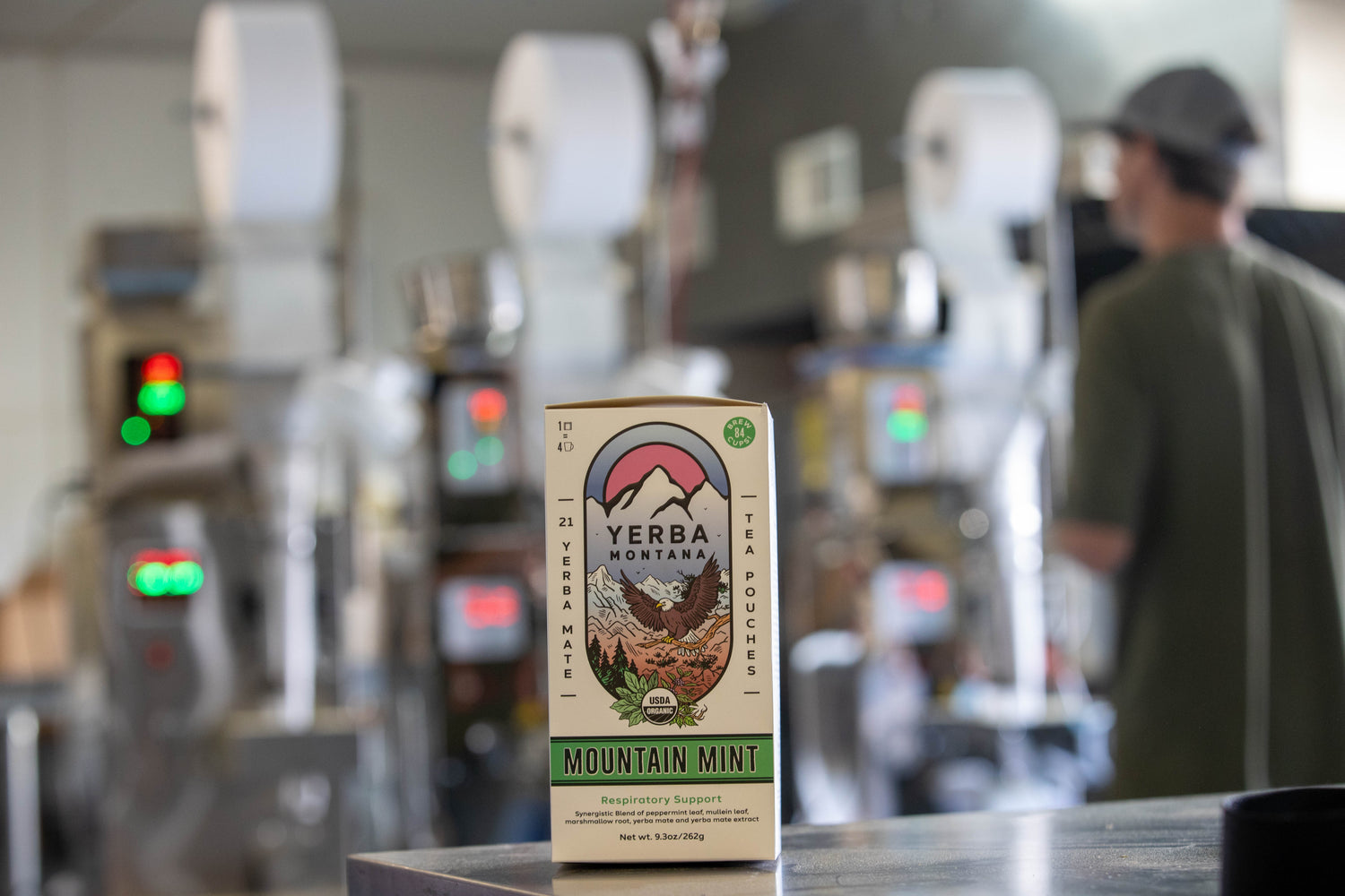 Yerba Montana Mountain Mint tea pouch box in focus, with blurry packaging gear and founder in the background

